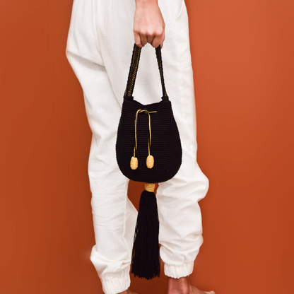 Mochila Tara Wayuu artesanal color negro, tejida a mano con hilos de algodón y seda dorada en el norte de Colombia. Mini mochila de diseño monocromático, con textura destacada, gasa corta, cordón ajustable y bailarina colgante. Fusión de tradición y elegancia contemporánea. Mochila Wayuu, mochila de fiesta, bolso wayuu, bolso de fiesta.