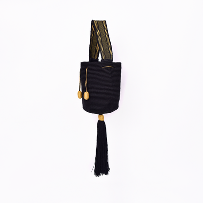 Mochila Tara Wayuu artesanal color negro, tejida a mano con hilos de algodón y seda dorada en el norte de Colombia. Mini mochila de diseño monocromático, con textura destacada, gasa corta, cordón ajustable y bailarina colgante. Fusión de tradición y elegancia contemporánea. Mochila Wayuu, mochila de fiesta, bolso wayuu, bolso de fiesta.